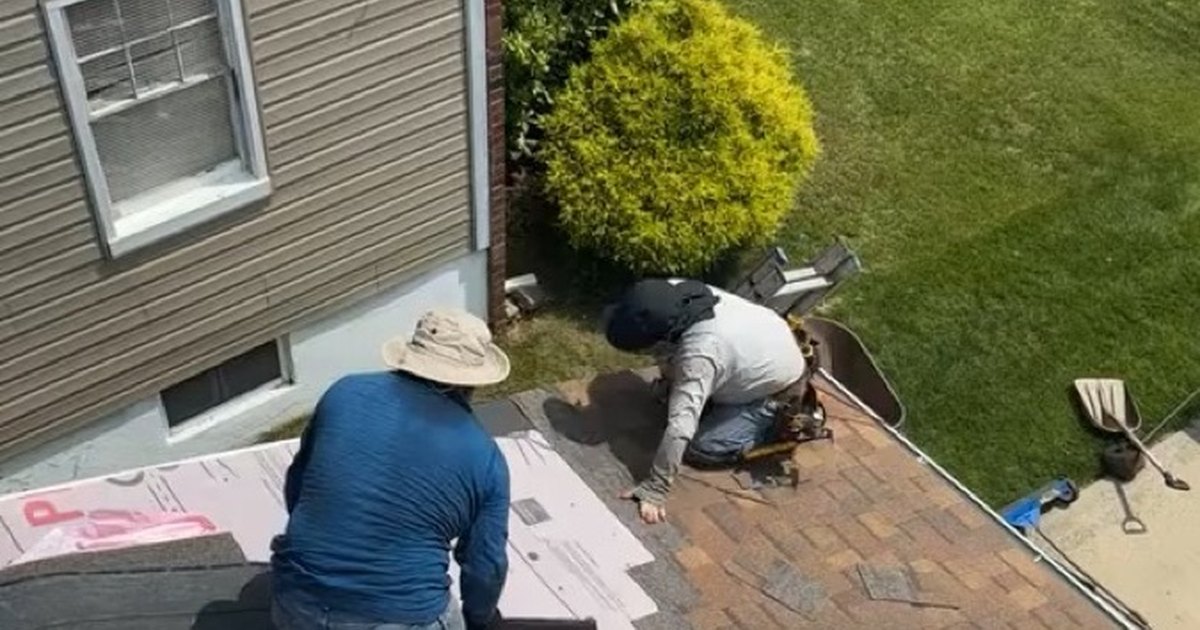 Two roofers laying architectural shingles over synthetic underlayment on a house beside a lawn