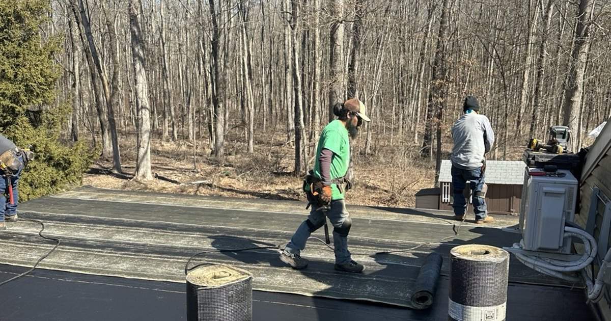 Workers and roofing rolls on a flat roof beside trees
