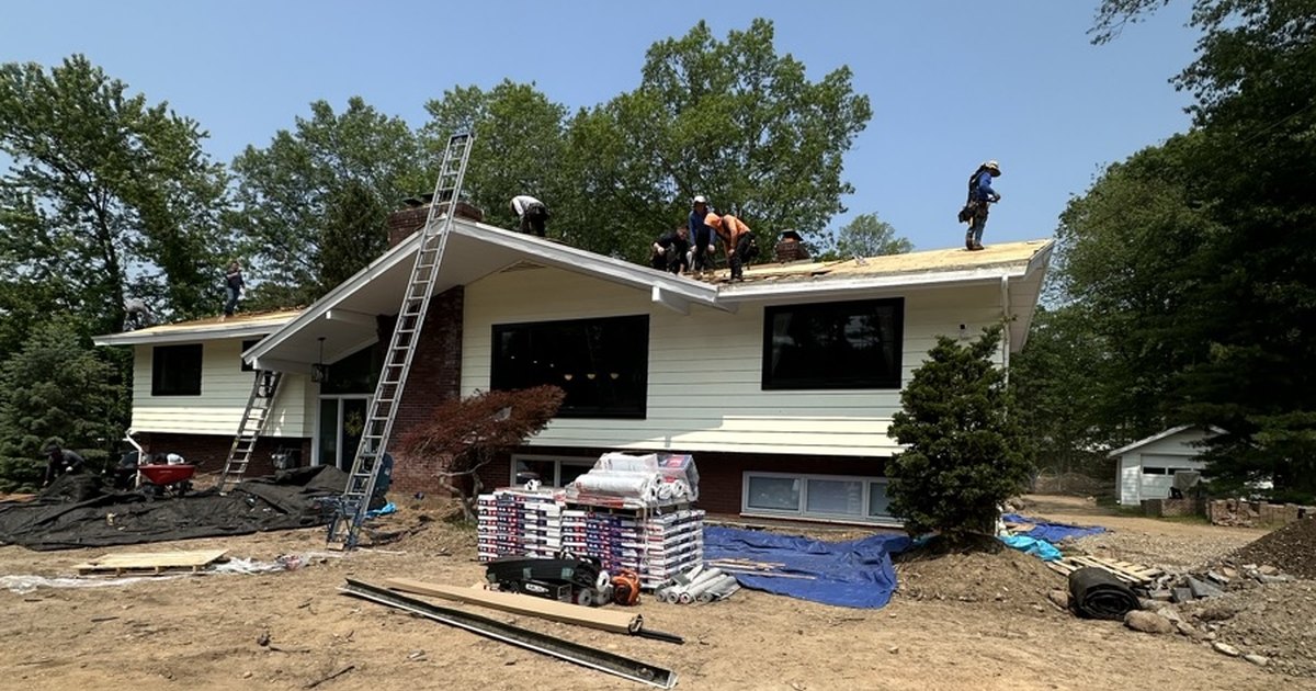 Crew working on bare roof decking during a tear-off, with shingle bundles staged in front of the house